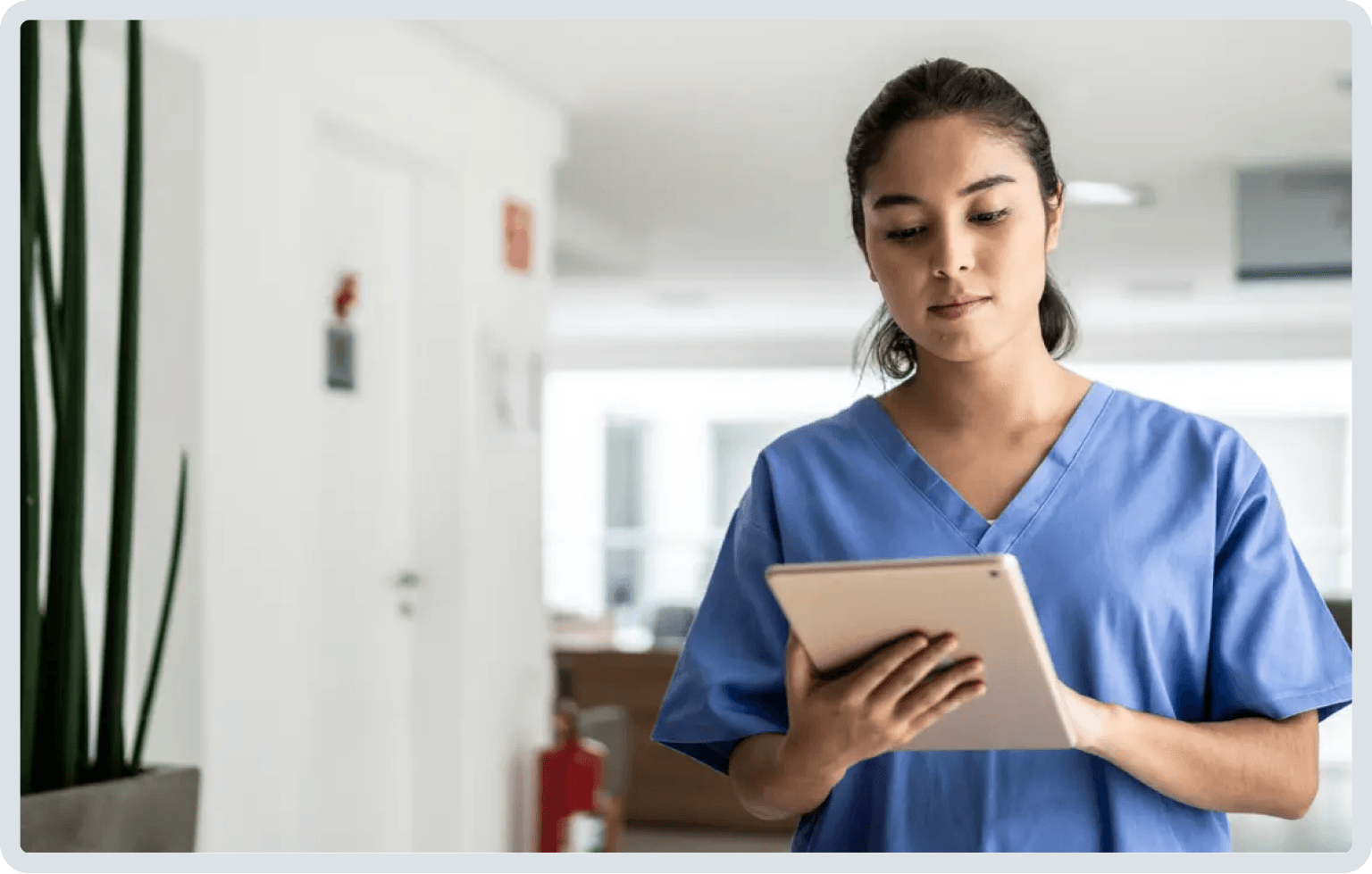 Femme en blouse bleue regardant une tablette dans un couloir.