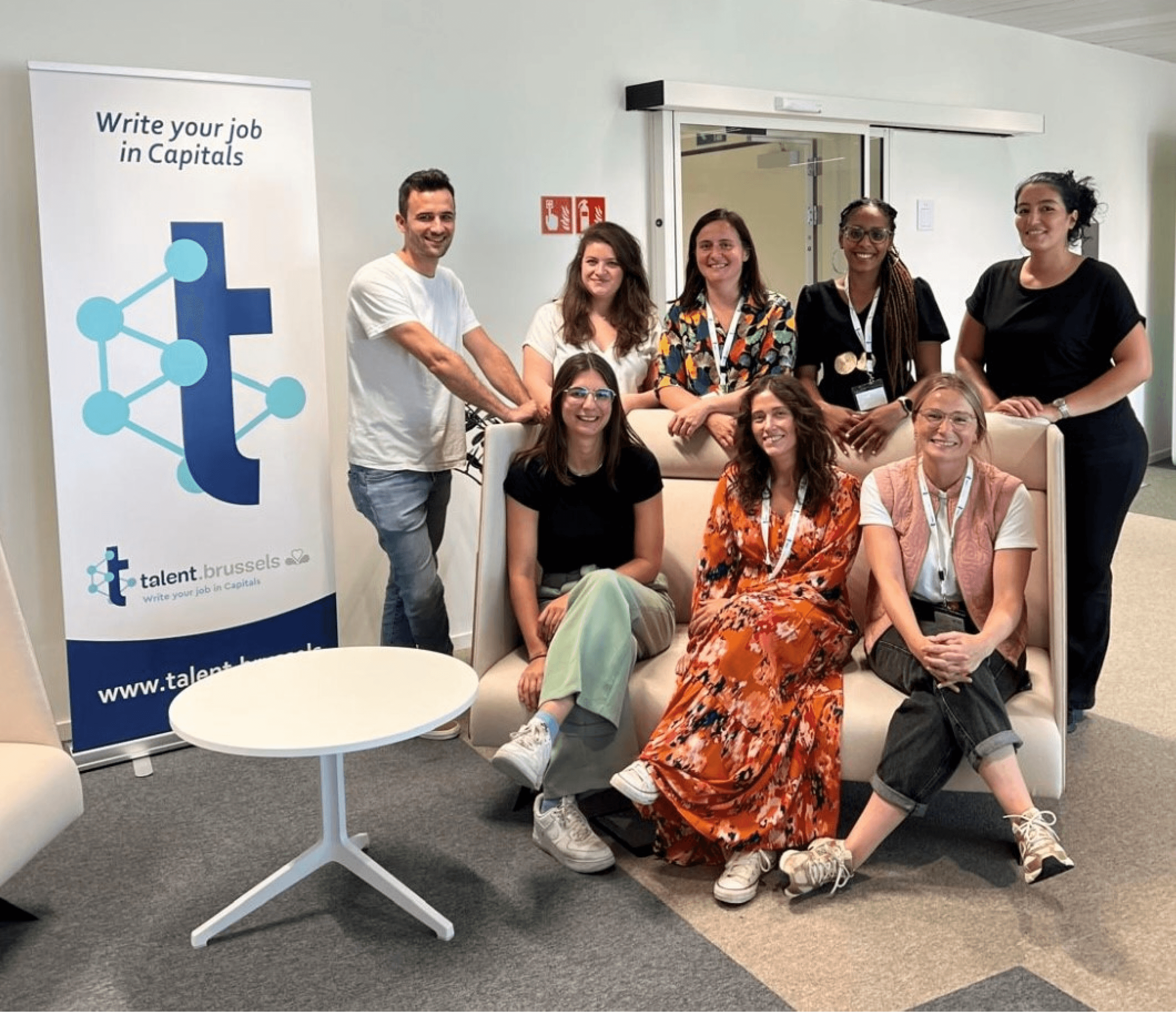 Eight smiling people, one man and seven women, pose in an office with a "talent.brussels" banner.